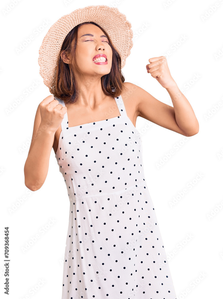 Young beautiful chinese girl wearing summer hat very happy and excited doing winner gesture with arms raised, smiling and screaming for success. celebration concept. Young beautiful chinese girl wearing summer hat very happy and excited doing winner gesture with arms raised, smiling and screaming for success. celebration concept.