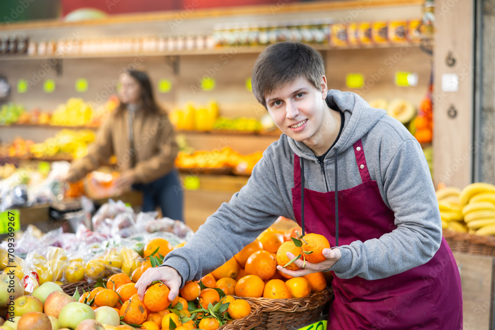 Attentive young salesman placing tangerines in a market basket in large grocery store