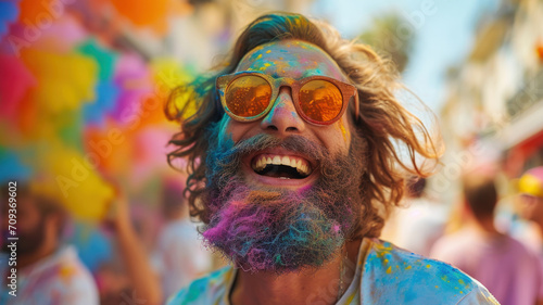 Happy man celebrating Holi festival, person with paint on face having fun. Portrait of bearded guy in sunglasses on colorful powder background. Concept of India, color, travel people