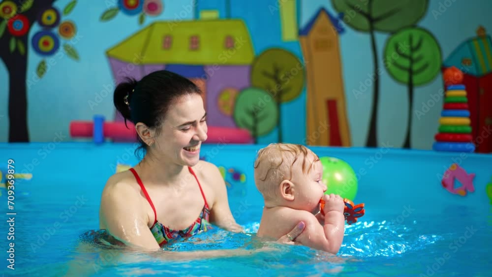 Smiling Caucasian female moving the infant in the swimming pool. Baby boy is holding a toy in his mouth. Smiling Caucasian female moving the infant in the swimming pool. Baby boy is holding a toy in his mouth.