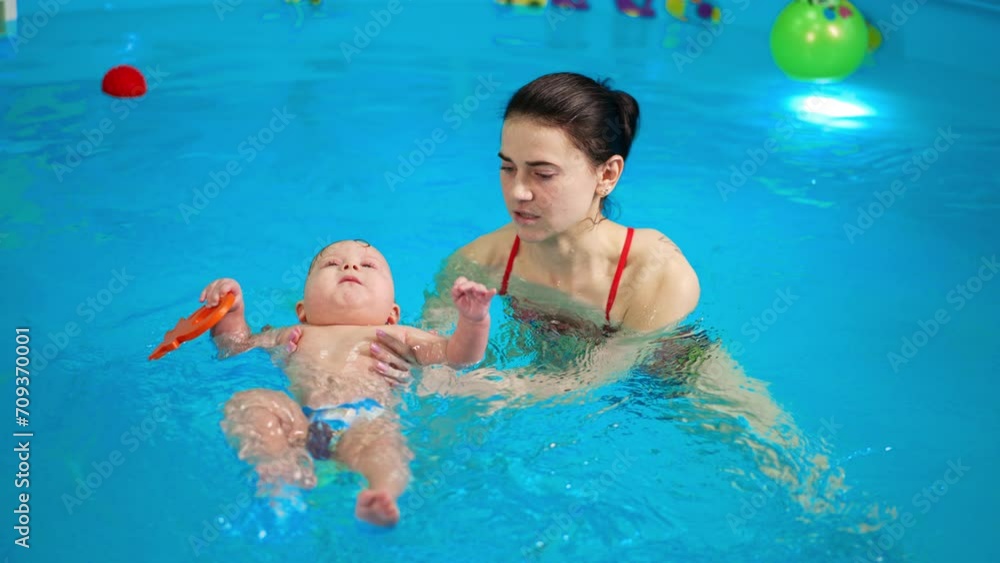 Sweet Caucasian infant baby lies on the water in the swimming pool. Female coach talking to a kid and moving him in water. Sweet Caucasian infant baby lies on the water in the swimming pool. Female coach talking to a kid and moving him in water.