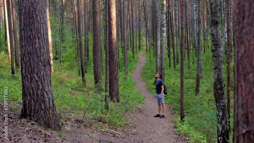 man in glasses, blue shorts and a gray T-shirt with a backpack walks through a pine forest, having rest outdoors at sunset.