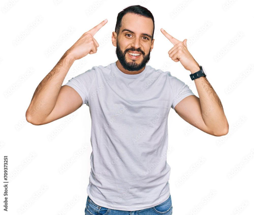 Young man with beard wearing casual white t shirt smiling pointing to head with both hands finger, great idea or thought, good memory Young man with beard wearing casual white t shirt smiling pointing to head with both hands finger, great idea or thought, good memory