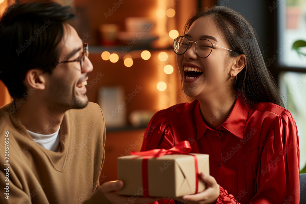 A joyful woman in glasses and a red shirt is laughing as she receives a gift from a man, also with glasses, in a warm indoor setting with soft lighting in the background. A joyful woman in glasses and a red shirt is laughing as she receives a gift from a man, also with glasses, in a warm indoor setting with soft lighting in the background.