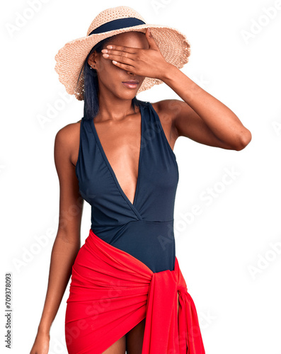 Young african american woman wearing swimsuit and summer hat covering eyes with hand, looking serious and sad. sightless, hiding and rejection concept