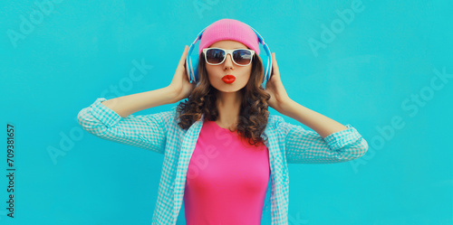 Portrait of stylish modern happy young woman listening to music with headphones wearing colorful pink hat on blue studio background