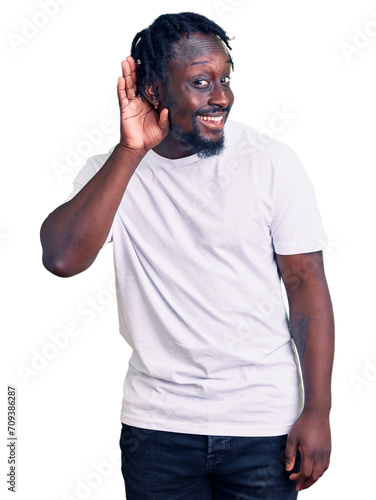 Young african american man with braids wearing casual white tshirt smiling with hand over ear listening an hearing to rumor or gossip. deafness concept.