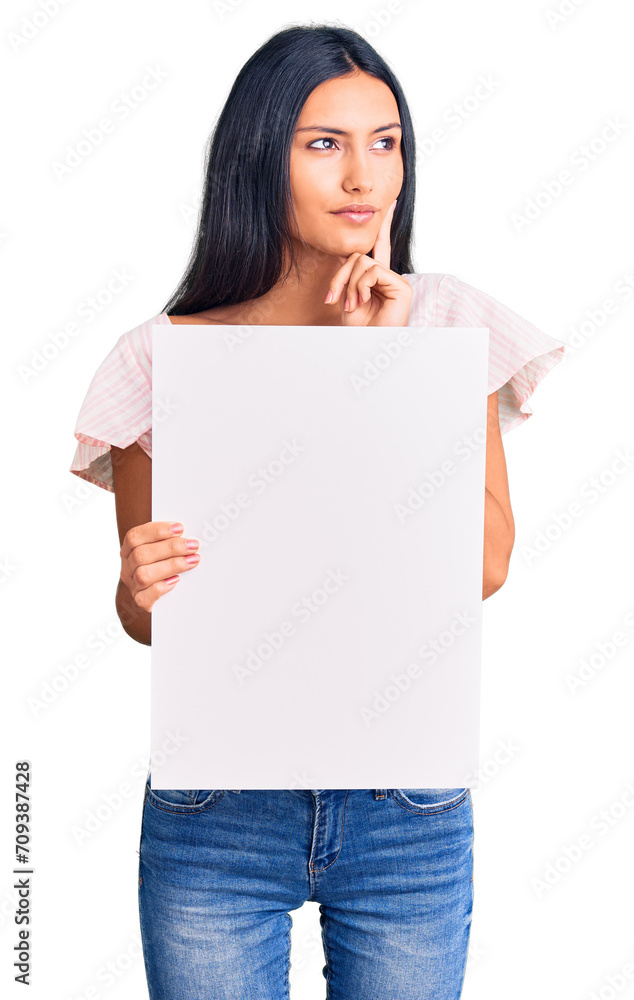 Young beautiful latin girl holding blank empty banner serious face thinking about question with hand on chin, thoughtful about confusing idea Young beautiful latin girl holding blank empty banner serious face thinking about question with hand on chin, thoughtful about confusing idea