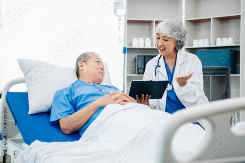 Asian doctor in white suit take notes while discussing and Asian elderly, man patient who lying on bed with receiving saline solution