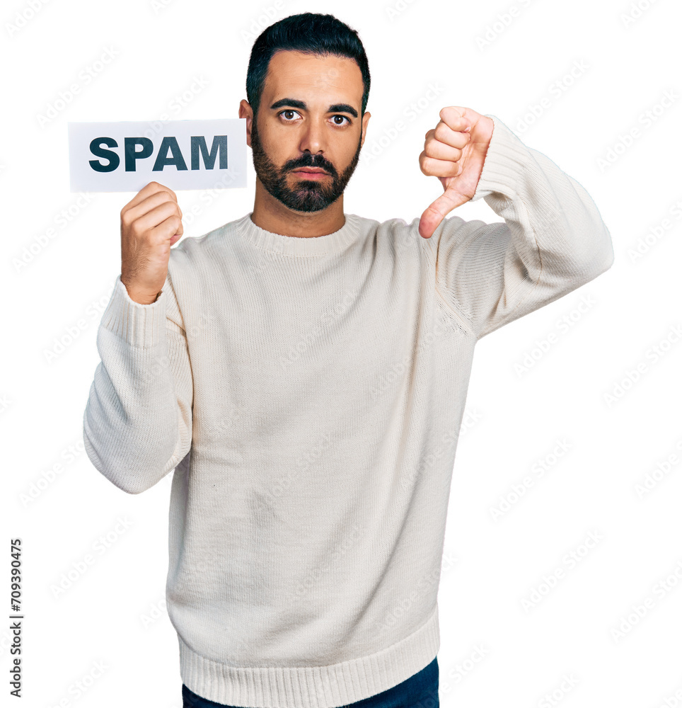 Young hispanic man with beard holding spam banner with angry face, negative sign showing dislike with thumbs down, rejection concept Young hispanic man with beard holding spam banner with angry face, negative sign showing dislike with thumbs down, rejection concept