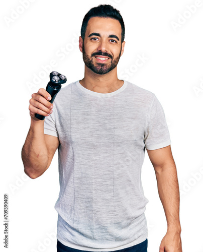 Young hispanic man with beard holding electric razor machine looking positive and happy standing and smiling with a confident smile showing teeth