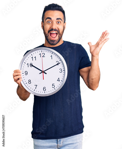 Young hispanic man holding big clock celebrating victory with happy smile and winner expression with raised hands