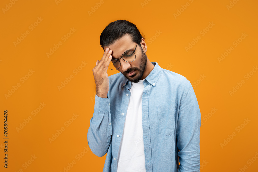 A pensive man in a light blue denim shirt touches his forehead, displaying a gesture of worry A pensive man in a light blue denim shirt touches his forehead, displaying a gesture of worry