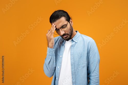 A pensive man in a light blue denim shirt touches his forehead, displaying a gesture of worry