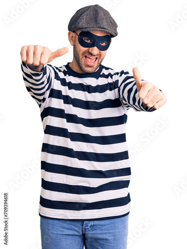 Young handsome man wearing burglar mask approving doing positive gesture with hand, thumbs up smiling and happy for success. winner gesture.