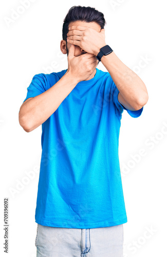 Young handsome man with beard wearing casual t-shirt covering eyes and mouth with hands, surprised and shocked. hiding emotion