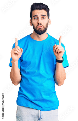 Young handsome man with beard wearing casual t-shirt pointing up looking sad and upset, indicating direction with fingers, unhappy and depressed.