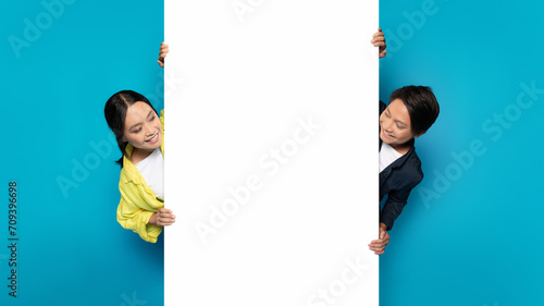 Asian man and woman playfully peeking around a large white vertical banner photo