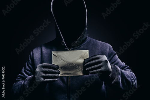 Photo of scary shaded hooded man holding paper letter on dark background.