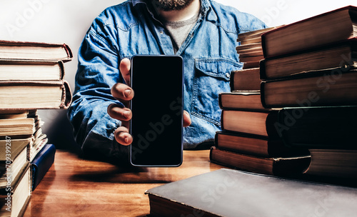 Photo of male person in denim shirt showing smartphone and sitting by the table with old book stacks, selling antiquities or audio books concept.