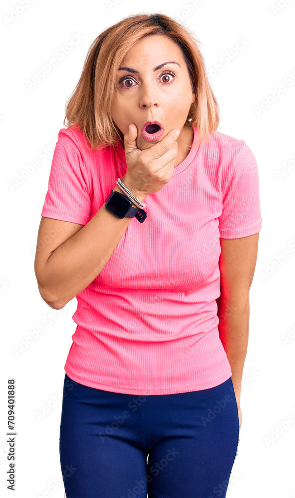 Young blonde woman wearing sportswear looking fascinated with disbelief, surprise and amazed expression with hands on chin Young blonde woman wearing sportswear looking fascinated with disbelief, surprise and amazed expression with hands on chin