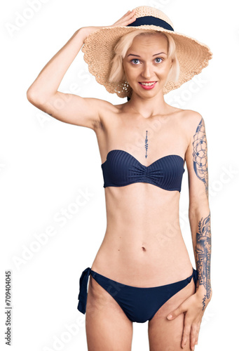 Young blonde woman with tattoo wearing bikini and summer hat confuse and wonder about question. uncertain with doubt, thinking with hand on head. pensive concept.