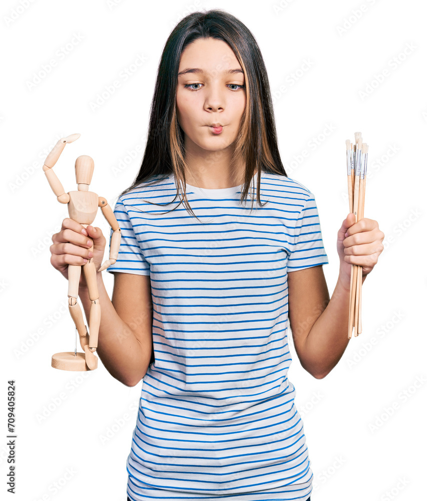 Young brunette girl with long hair holding small wooden manikin and pencils making fish face with mouth and squinting eyes, crazy and comical.