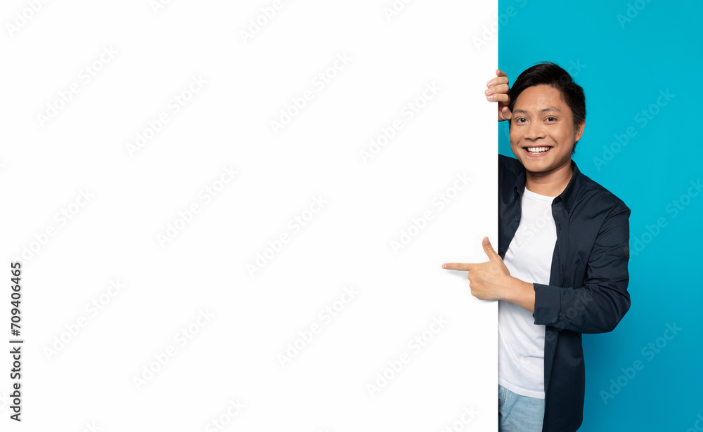 Happy Asian man in casual attire peeking around a large blank white banner Happy Asian man in casual attire peeking around a large blank white banner