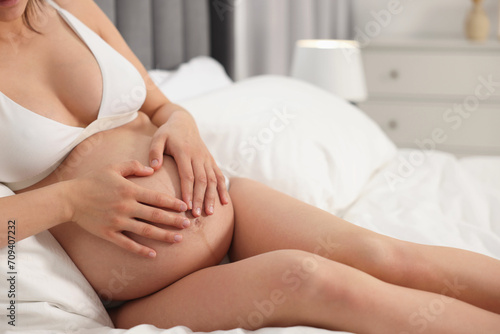 Pregnant woman in stylish comfortable underwear making heart with hands on her belly in bedroom, closeup