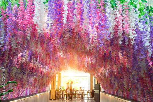 Artificial pink and violet wisteria flowers arch. Spring floral installation scene with geometric arch form. Wedding round tunnel with sunlight at the end of the hole.
