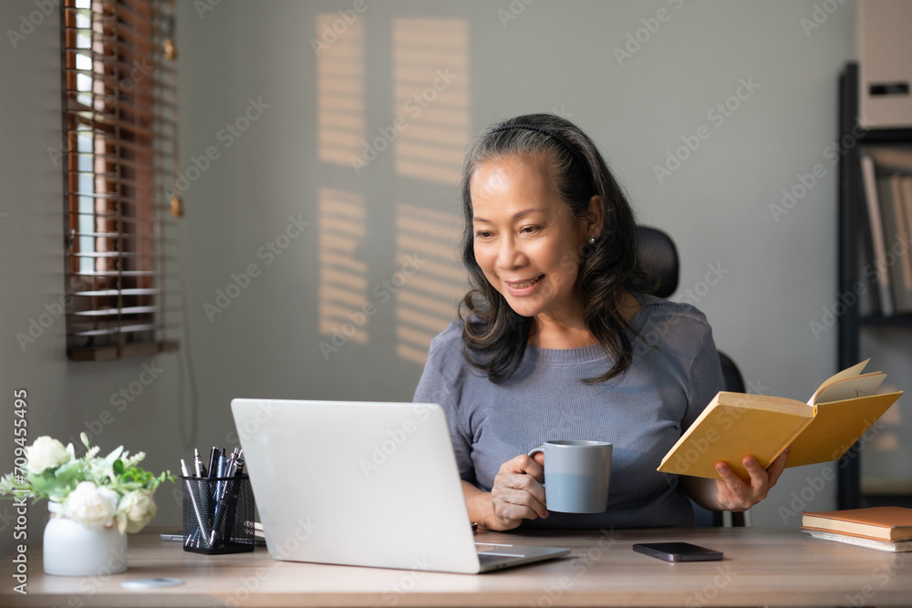 Senior asian woman holding coffee and book with laptop computer at home. Senior asian woman holding coffee and book with laptop computer at home.