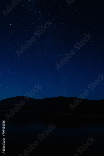 Dark blue starry sky, mountain silhouette, and tranquil lake at night.