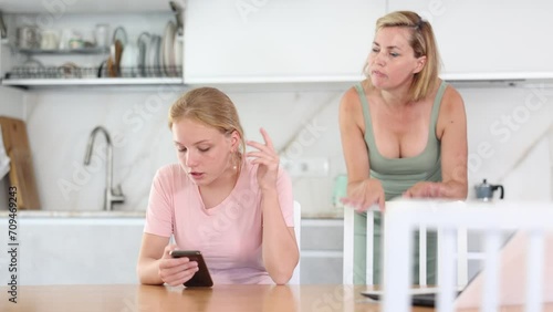 Upset teenage girl immersed in phone sitting at home table while disapproving mother standing in background, reflecting concern over daughter detachment from environment  photo