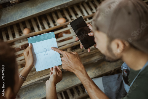 Asian male worker checks savings book when using mobile phone at chicken farm