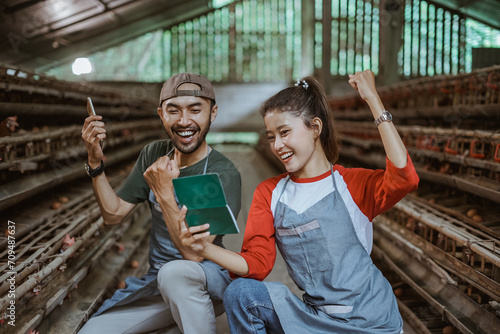 Happy male and female workers holding saving book with clenched hands up at a chicken farm