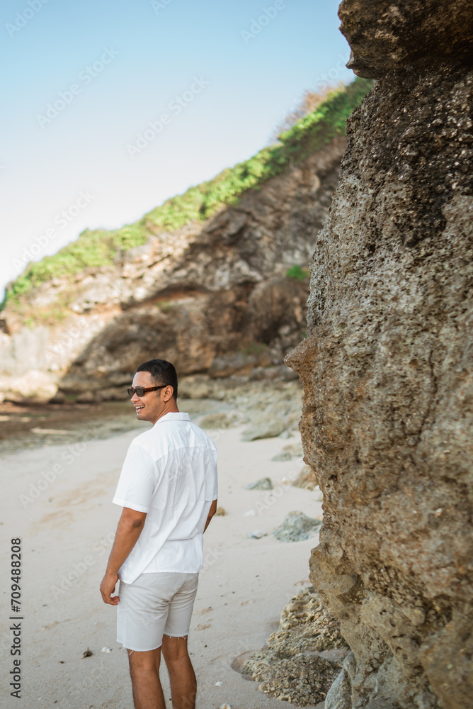 Fototapeta premium happy asian man in white with sunglasses standing next to cliff on exotic tropical beach