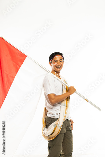 a handsome man in white t shirt and sarong as his shoulder belt standing bring a stick with the indonesian flag on it and smiling