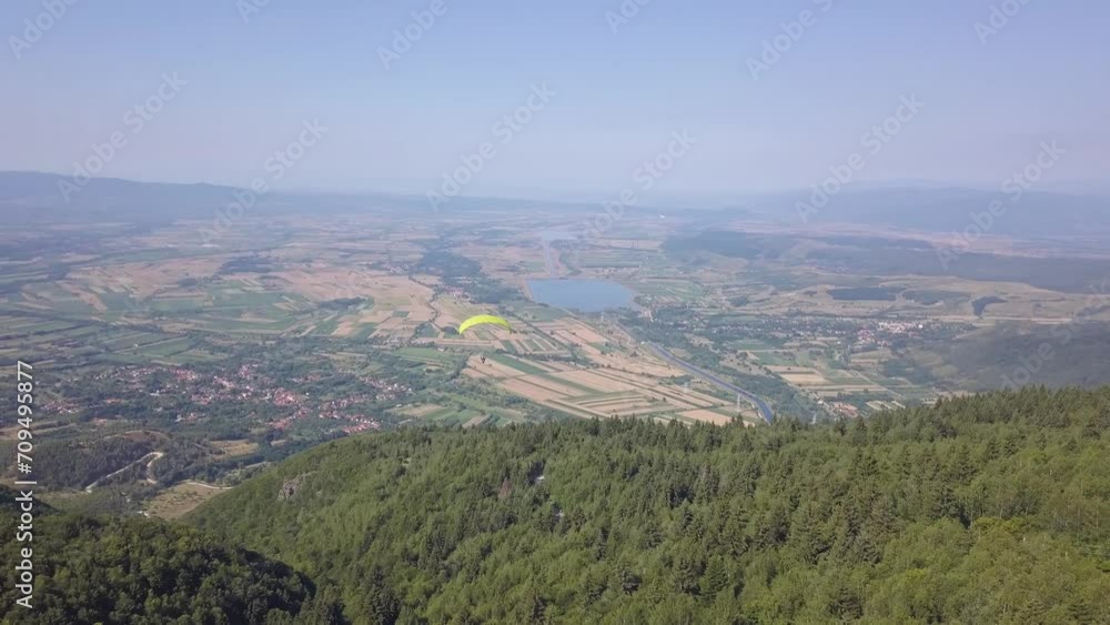 Aerial view from drone of paragliding. Person in far distance, under large yellow parachute fly high over mountain terrain soaring through the air. Aerial view from drone of paragliding. Person in far distance, under large yellow parachute fly high over mountain terrain soaring through the air.