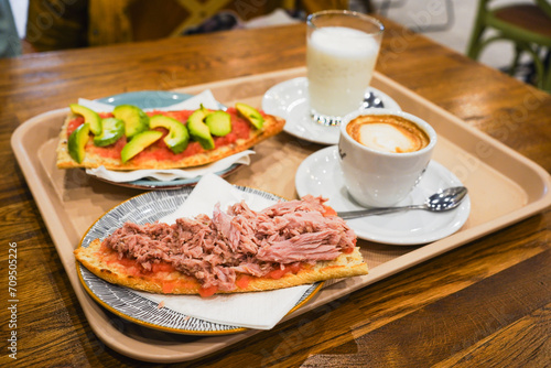 Breakfast tray with avocado toast, tuna on toast, coffee, and milk on a cafe table.