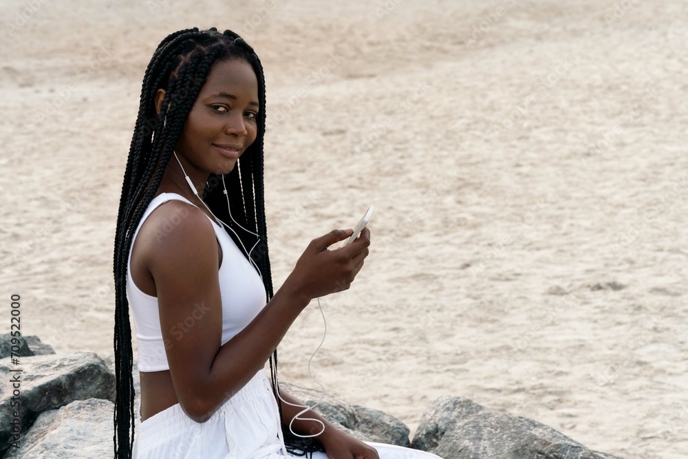 Woman with braids with headphones sitting at the beach using phone
