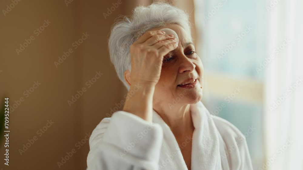 Senior woman look at mirror touch face. Wellbeing smiling woman in bathrobe with gray hair looking at mirror taking care of her face. Getting old age change, face skincare self-care concept. Senior woman look at mirror touch face. Wellbeing smiling woman in bathrobe with gray hair looking at mirror taking care of her face. Getting old age change, face skincare self-care concept.