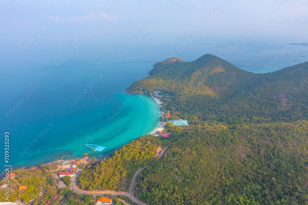 Tropical island with a sandy azure bay and pier and a beach village settlement surrounded with roads by exotic green forests trees hills, aerial view