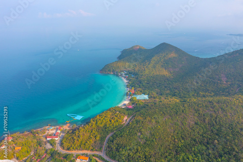 Tropical island with a sandy azure bay and pier and a beach village settlement surrounded with roads by exotic green forests trees hills, aerial view