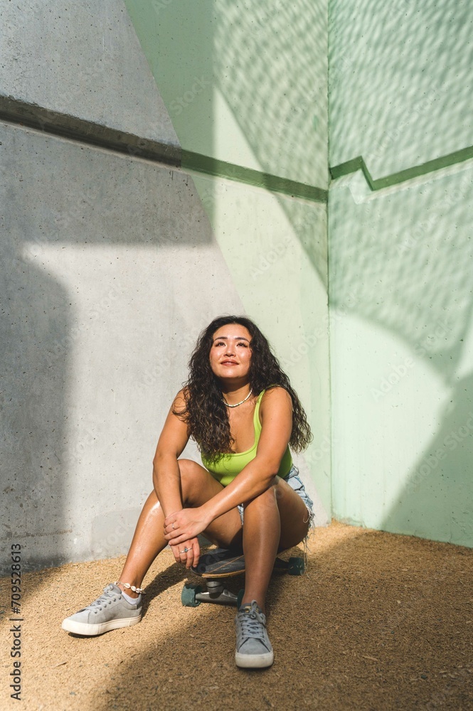 Asian woman sitting on her skateboard next to the  green wall