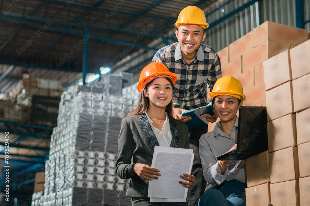 three factory employees smile at the camera while working on a laptop in the goods warehouse three factory employees smile at the camera while working on a laptop in the goods warehouse