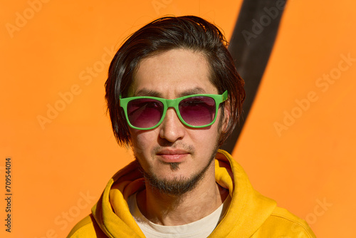Stylish Asian guy in green glasses stands against a yellow wall,