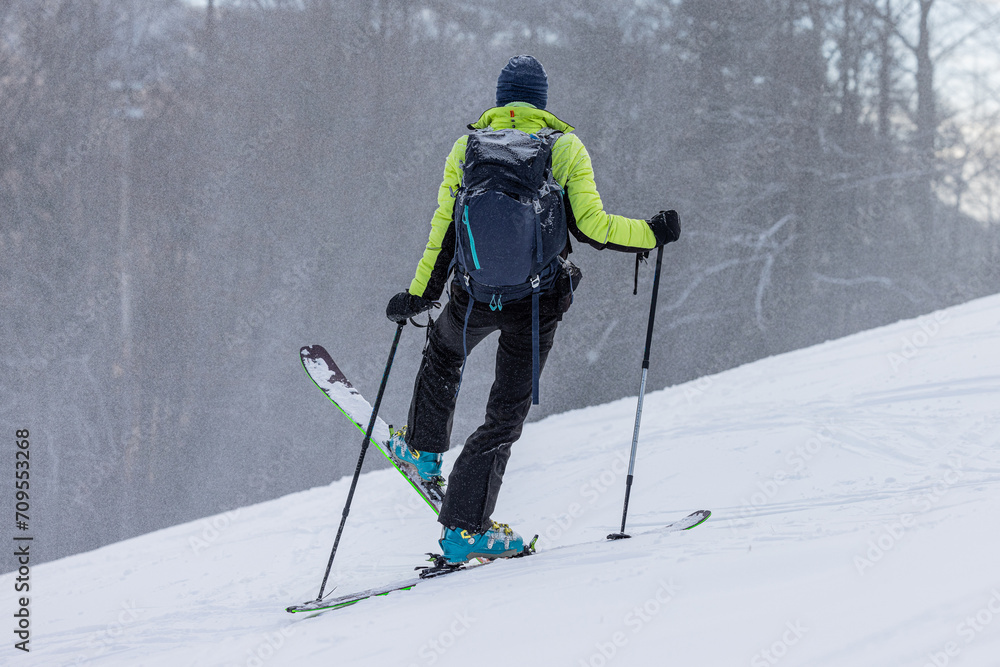 Ski touring, a person is performing a 45 degree turn while walking uphill with skis, modern equipment for ski touring, snow is falling down, trees in the background.