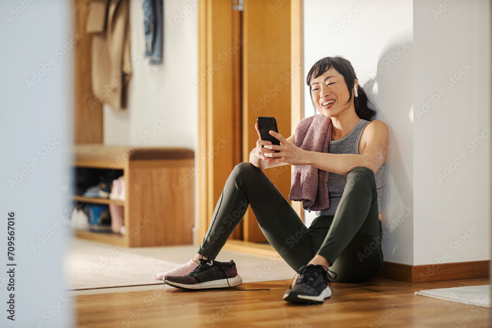 A fit middle-aged japanese woman is resting on the floor at home after workouts and looking at her fitness app.
