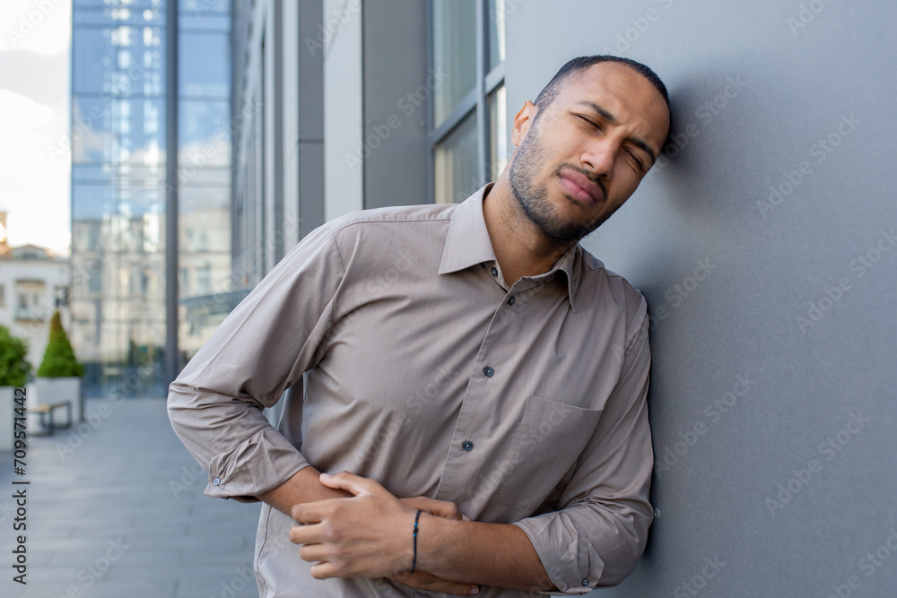 Man in pain outdoors, clutching his side and grimacing, conveying a sense of discomfort or abdominal ache against an urban backdrop. Man in pain outdoors, clutching his side and grimacing, conveying a sense of discomfort or abdominal ache against an urban backdrop.