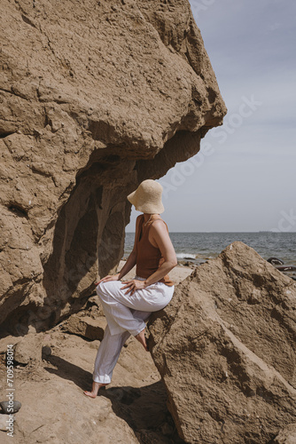 Woman in white pants and straw hat sitting on the rock and looking at sea. Elegant fashion, holidays, travel, vacation composition